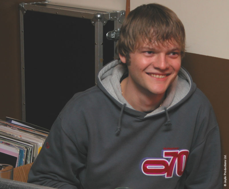 A smiling person in a grey hooded top sits in front of crates of records