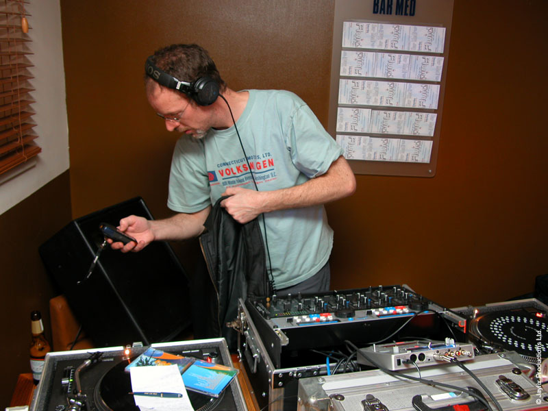 A DJ in headphones looks at a small device beside a mixer and turntables, a Bar Med sign on the wall behind