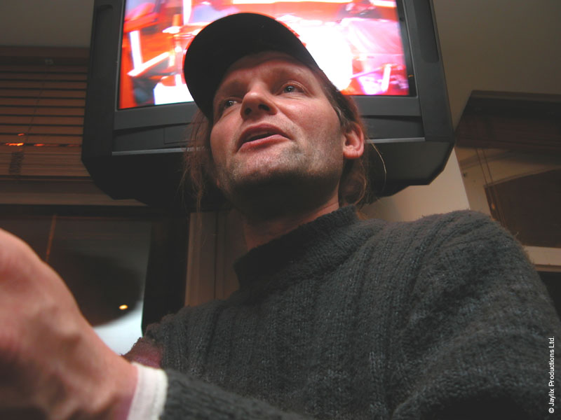 A man in a baseball cap and dark jumper looks up beside a television showing a bright picture