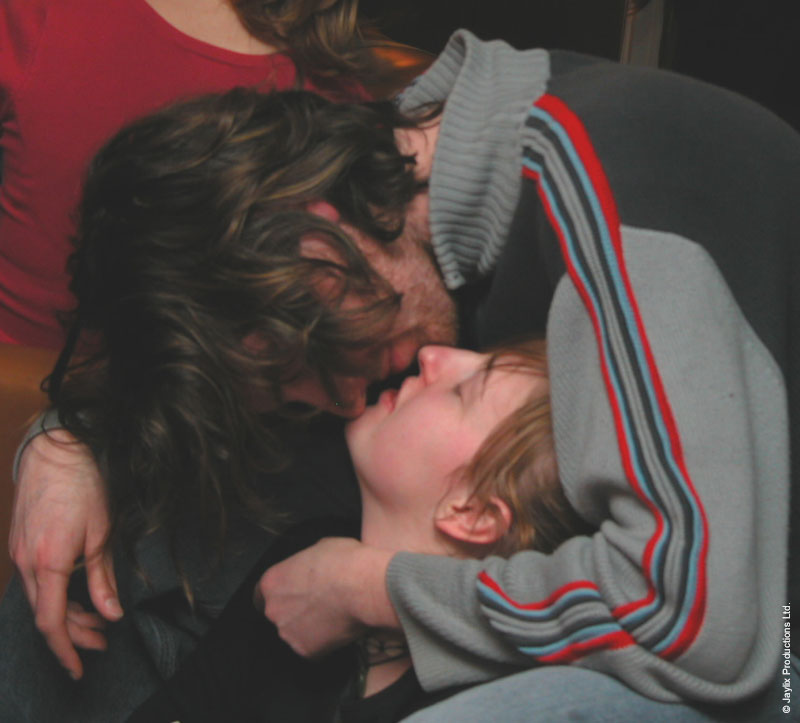 Two people embrace and kiss, one wearing a grey top with red and blue stripes on the sleeve