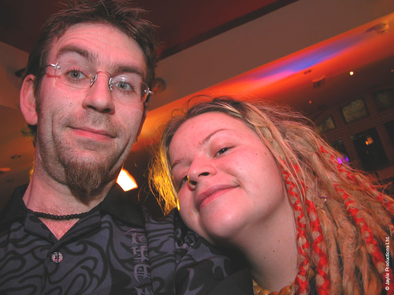 A man with glasses and a goatee and a person with red and blonde dreadlocks lean together and smile