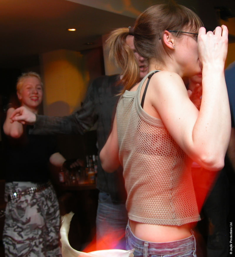 A woman in a mesh top and glasses dances with her hand at her face, with a person smiling in the background