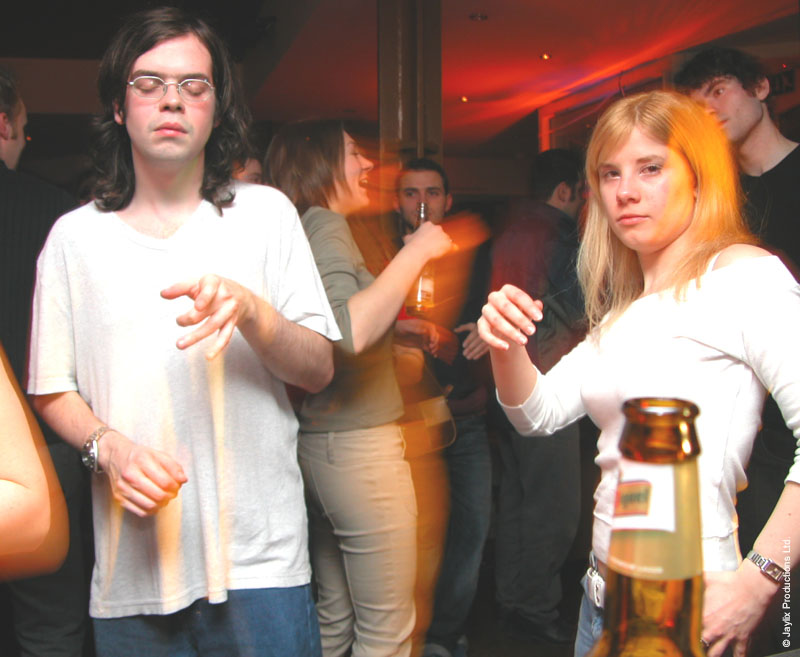 A person in a white t-shirt holding drumsticks and a woman in white holding a bottle stand in a busy bar