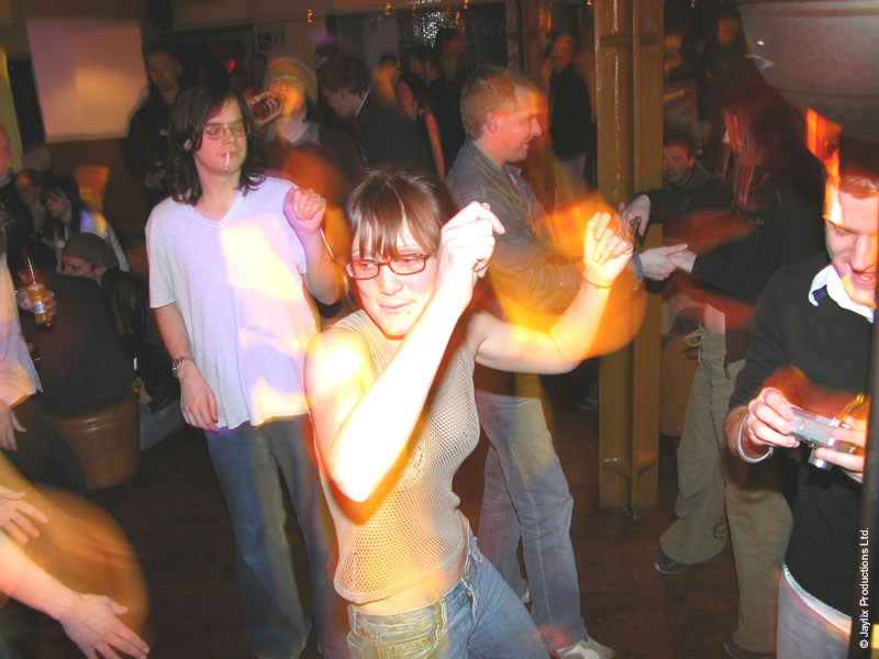 A woman with glasses dances in a crowded room while others look on, some holding cameras
