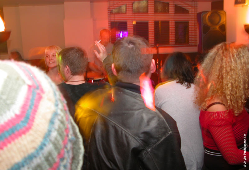 A crowd seen from behind in a bar, including a person in a striped woolly hat and a person in a leather jacket