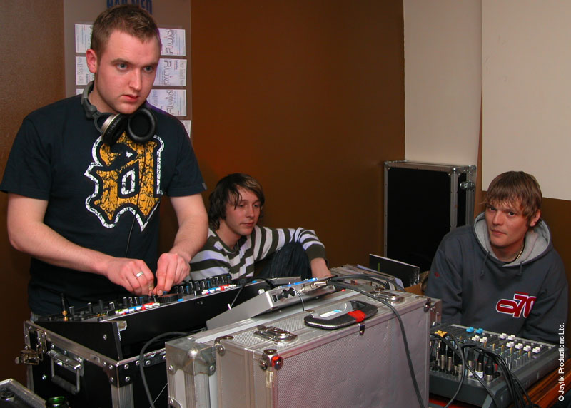 A DJ in a dark t-shirt mixes at a decks setup while two people sit behind