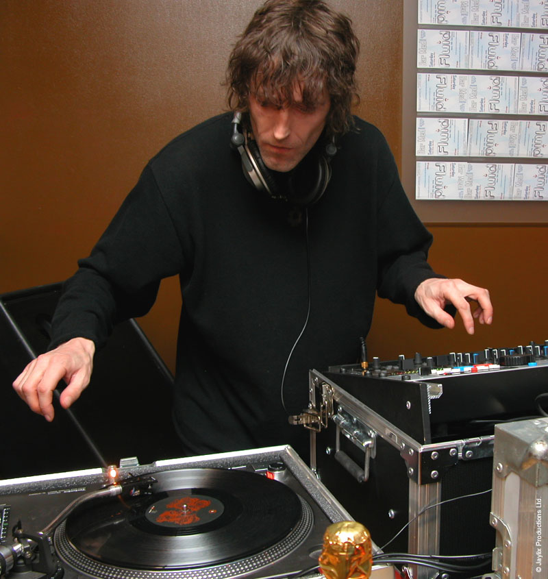 A DJ with headphones around his neck reaches toward a record on a turntable, with flyers on the wall behind