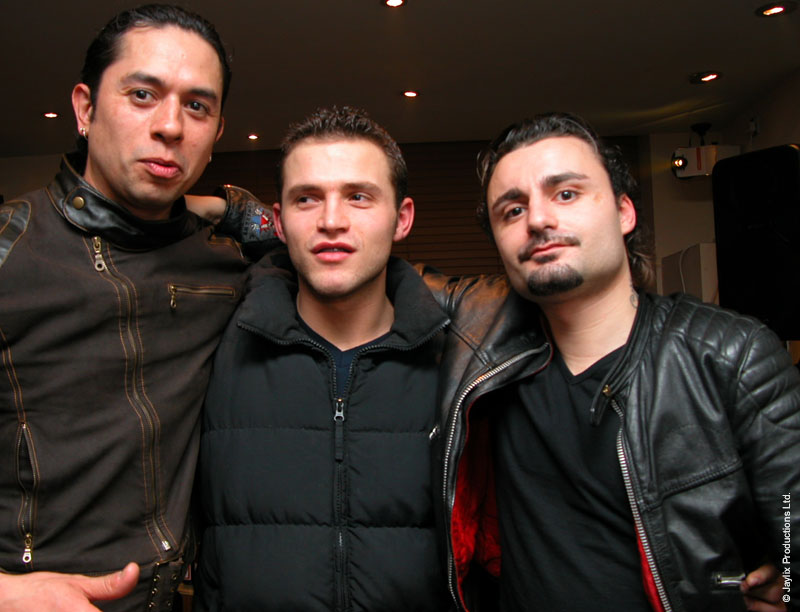 Three men in jackets pose with their arms around each other in a bar