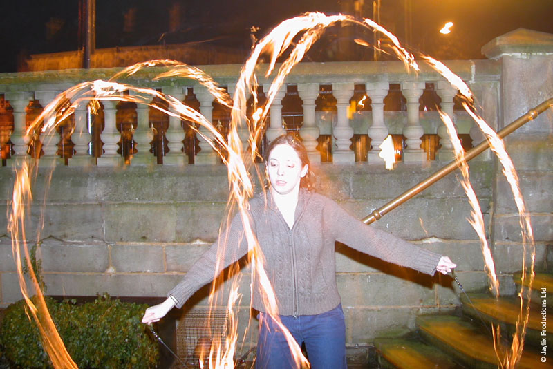 A person stands with arms outstretched in front of a stone balustrade, surrounded by trails of fire from poi