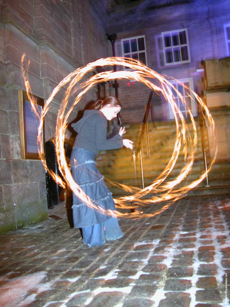 A person in a long denim skirt spins fire poi on a snowy cobbled street outside a stone building