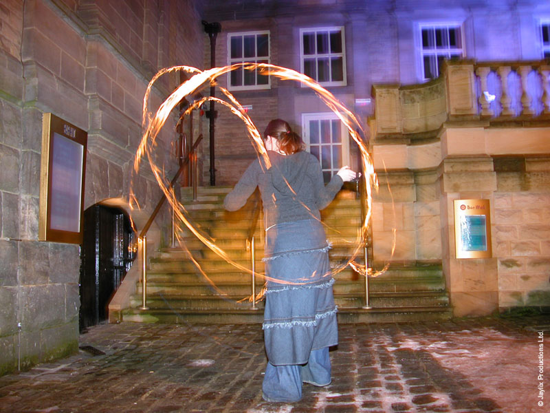 A person in a long denim skirt spins fire poi in front of stone steps and a doorway at night