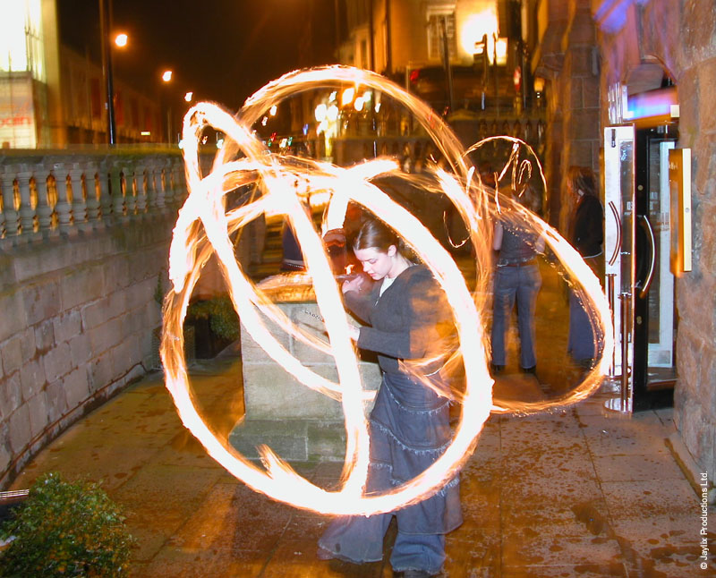 A person in a long denim skirt spins fire poi on a street, with overlapping trails of light