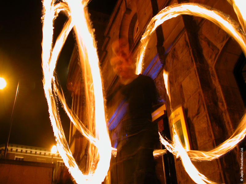 Streaks of fire from spinning poi in front of a dark stone building at night, with a blurred figure