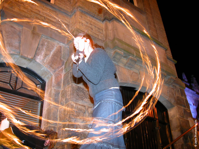 A person in a denim skirt spins fire poi against a stone building, surrounded by circular trails of flame