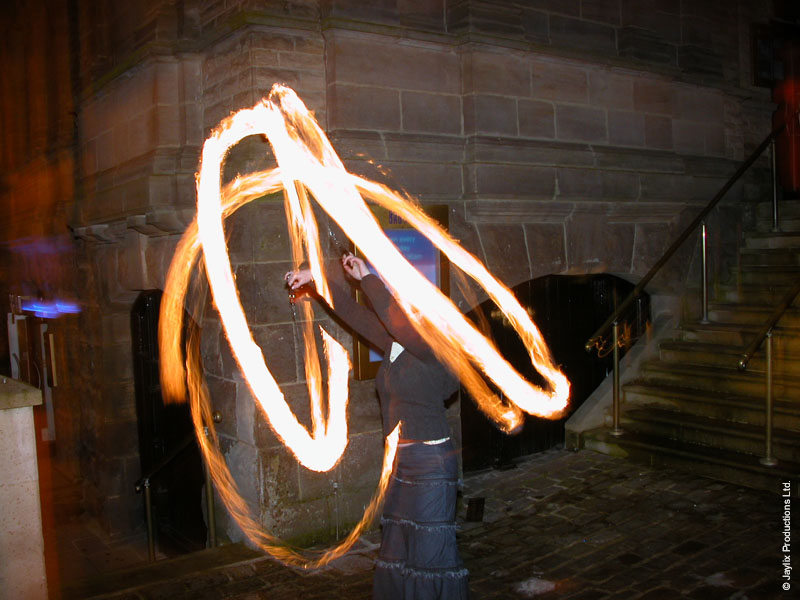 A person spins fire poi beside a stone building at night, leaving looping light trails