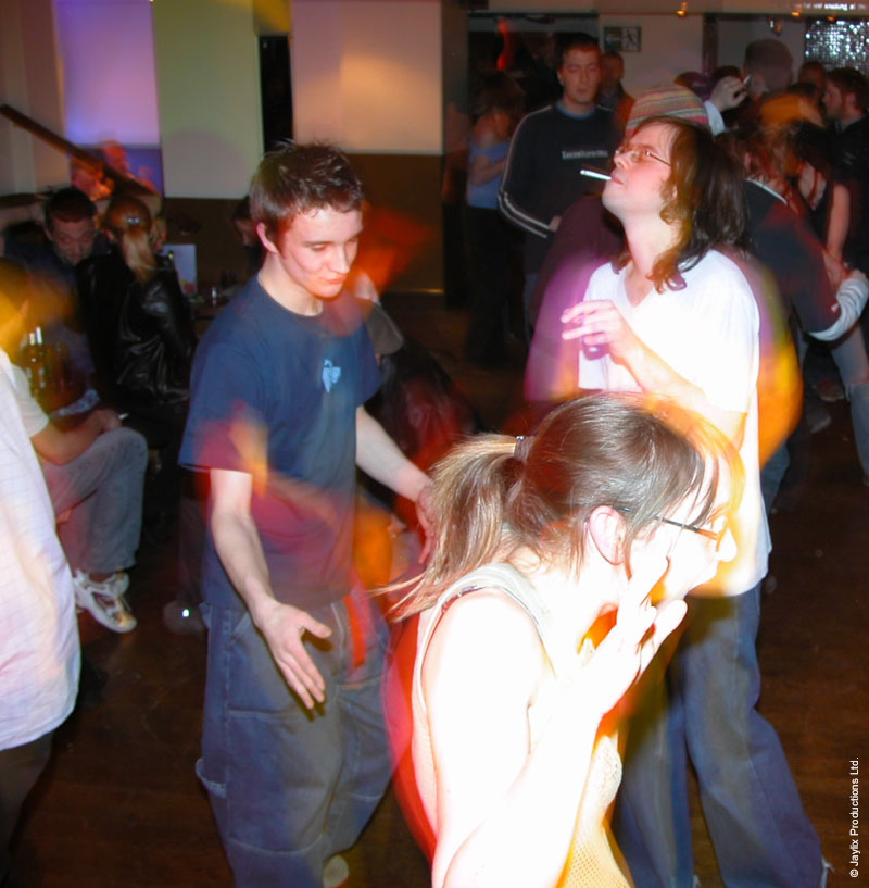 People dance in a dim bar, a man in a blue t-shirt in the foreground and a woman with glasses in front