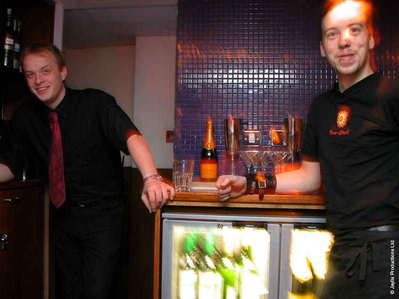 Two smiling men in black shirts stand behind a bar counter with a bottle of champagne and a fridge of drinks