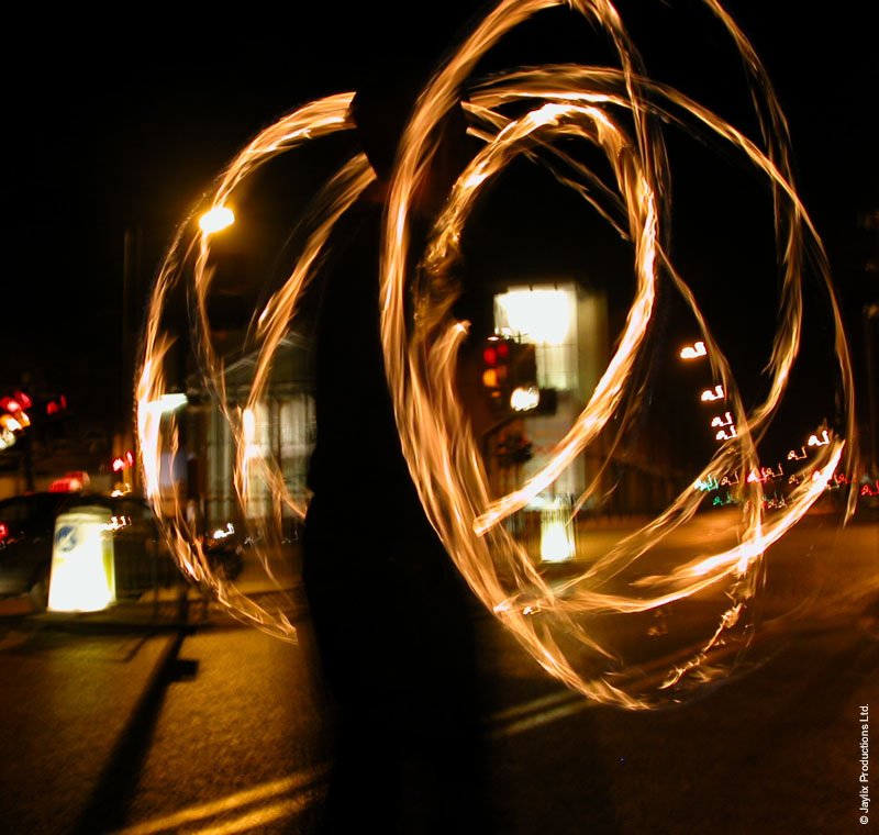 Looping trails of fire from poi at night beside a street, with a dark silhouette