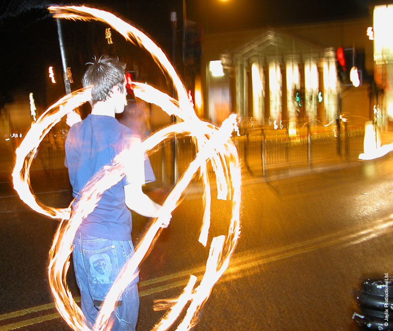 A person in a blue t-shirt spins fire poi on a road at night, with lit buildings behind