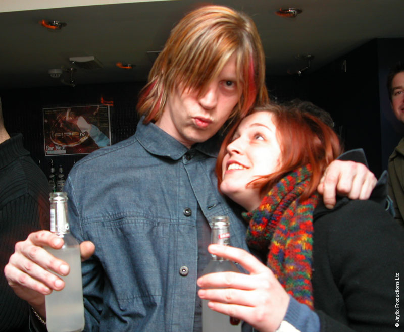 A person with a fringe and a denim shirt and a person with a colourful scarf hold bottles and pose together