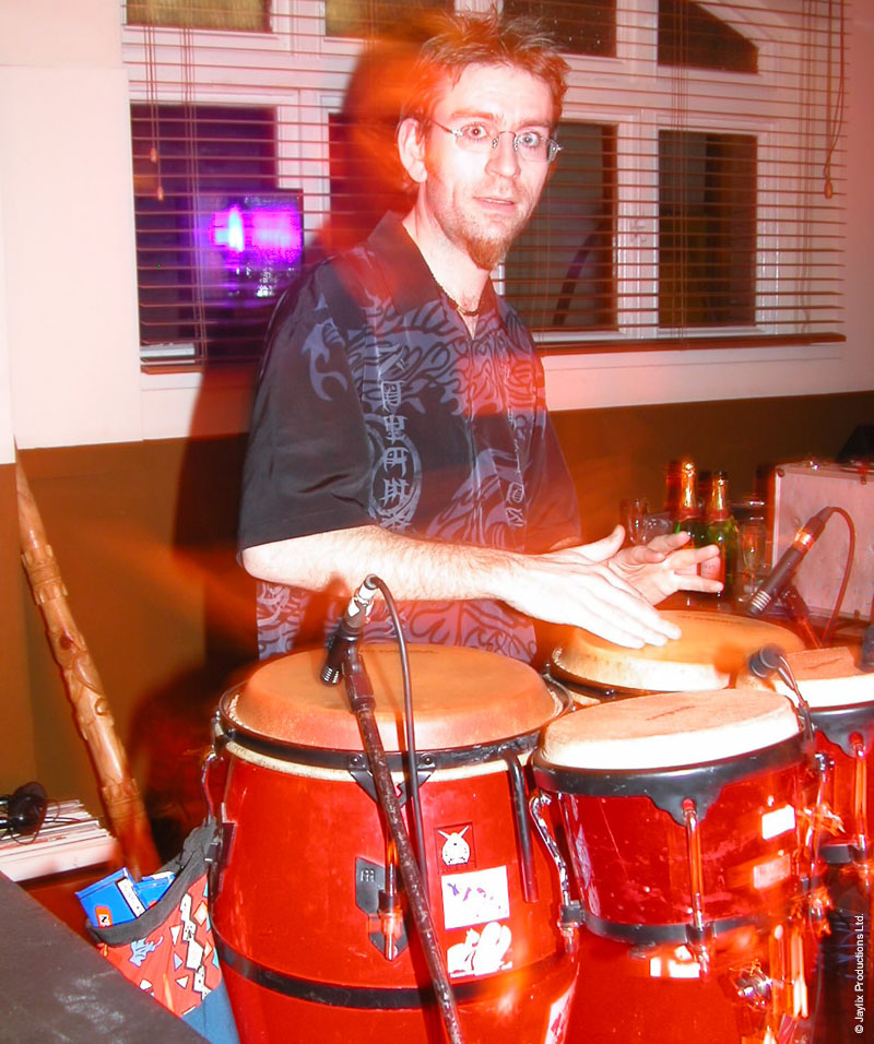 A man with glasses and a goatee plays red congas, with a window behind him