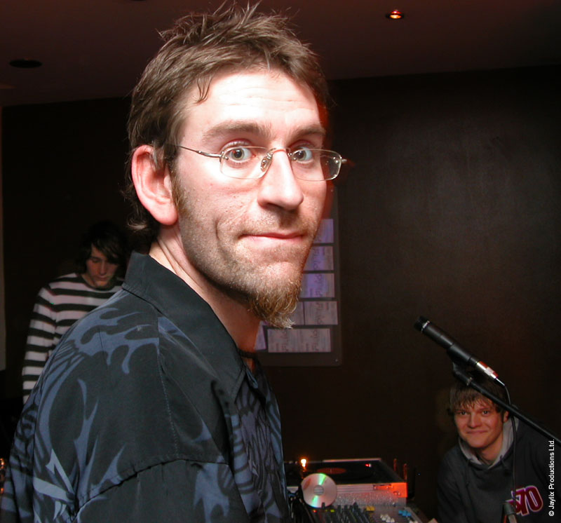 A man with glasses and a goatee looks at the camera, with a person in a striped top behind him