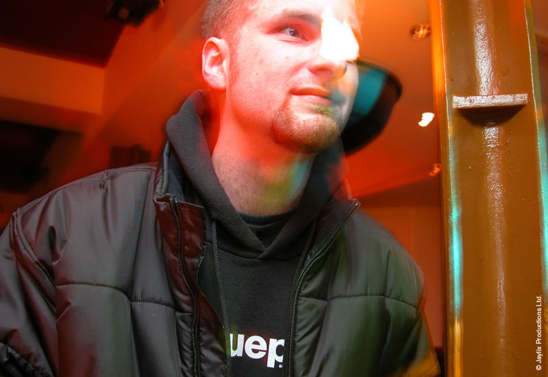 A man in a dark padded jacket and hooded top stands beside a yellow pillar, lit in orange