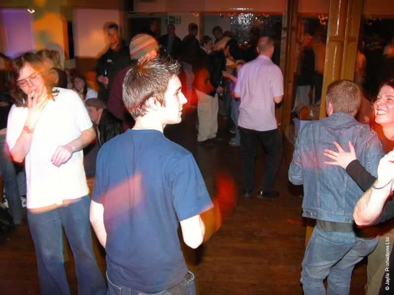 People dance in a wooden floored bar, a person in a blue t-shirt seen from behind in the centre