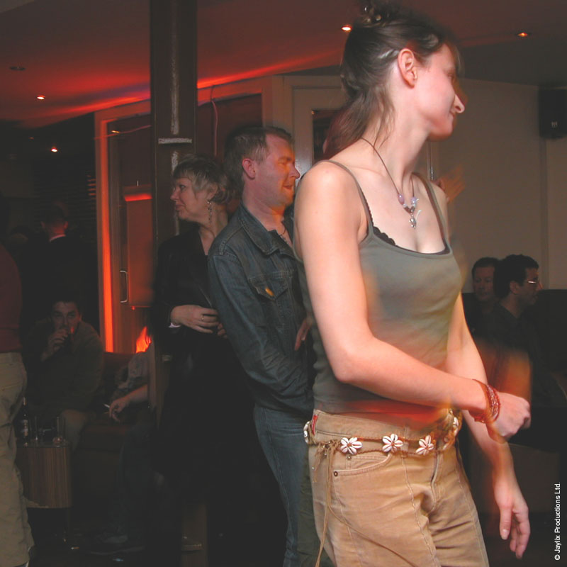 A woman in a grey vest top and tan trousers walks past in a dim bar while others stand behind