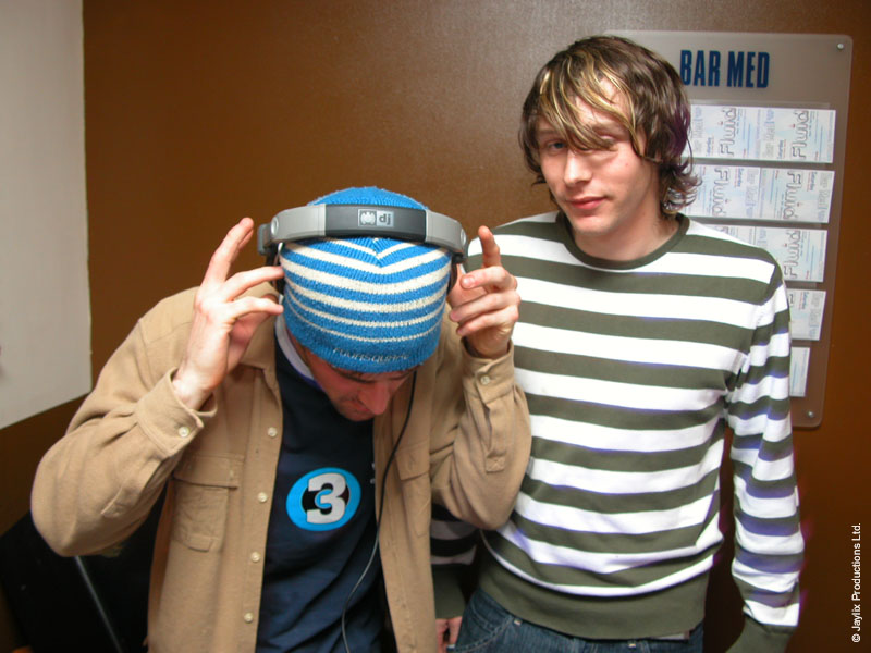 A person in a blue striped beanie holds headphones to their head beside a person in a striped top, in front of a Bar Med sign