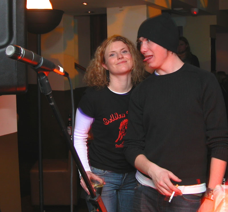 A woman in a black t-shirt and a person in a black beanie and jumper with a cigarette stand by a microphone stand