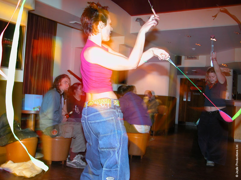 A person in a pink top and baggy jeans swings long ribbons in a bar, with another person behind