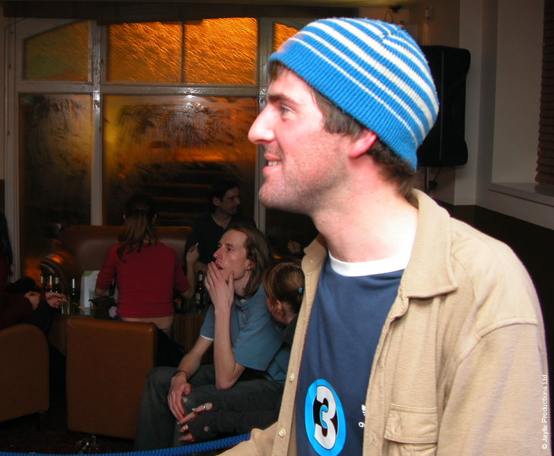 A smiling person in a blue striped beanie and beige jacket looks to the side, with seated people behind
