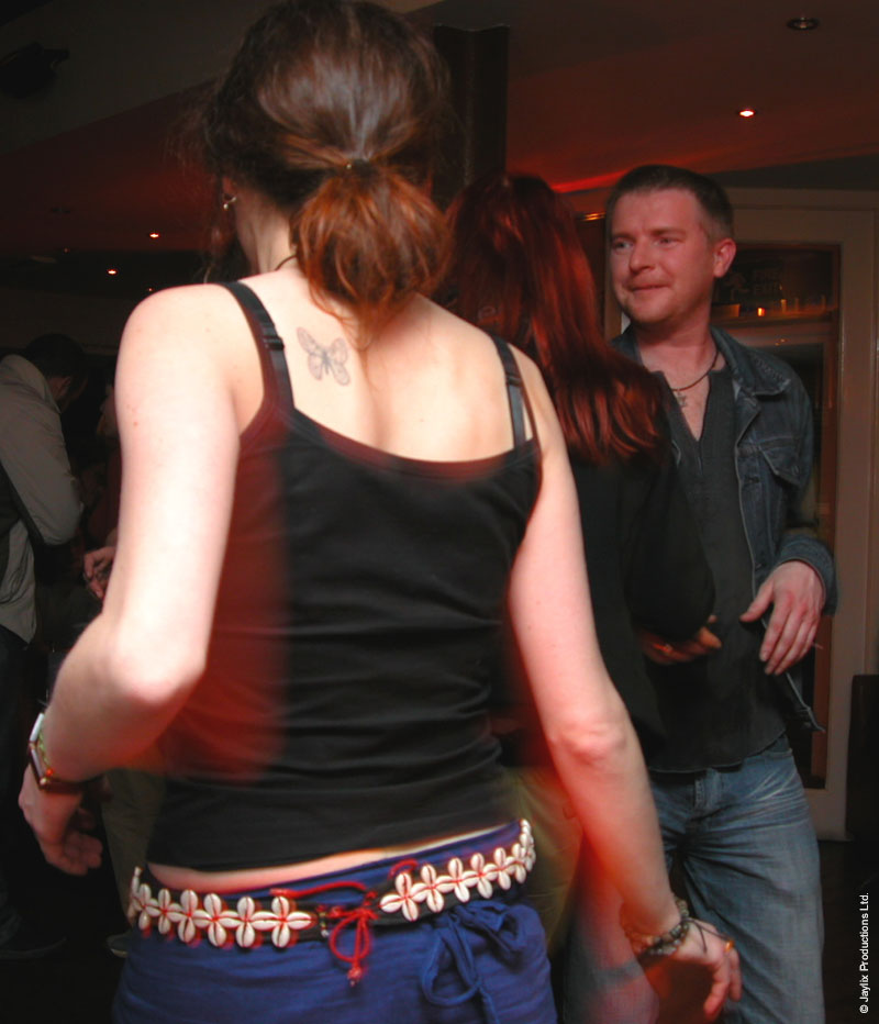 A woman in a black vest top with a tattoo on her back dances, with a man in a denim jacket smiling behind
