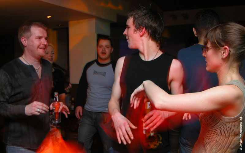 People stand and dance in a dim bar, with a woman in a mesh top on the right