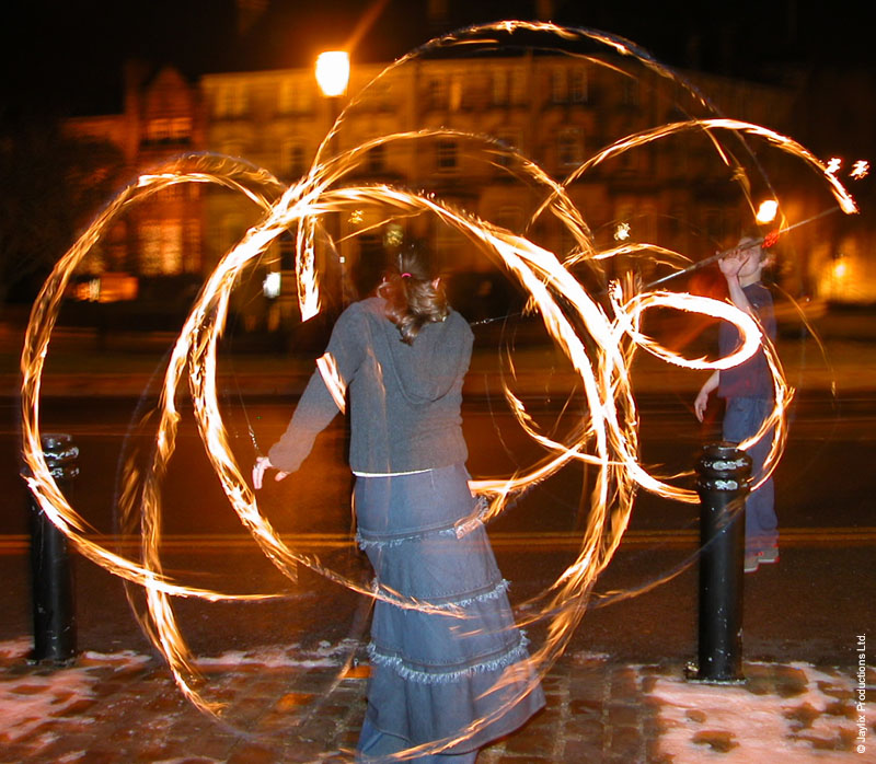 A person in a denim top and long skirt spins fire poi on a street at night, with a street lamp behind