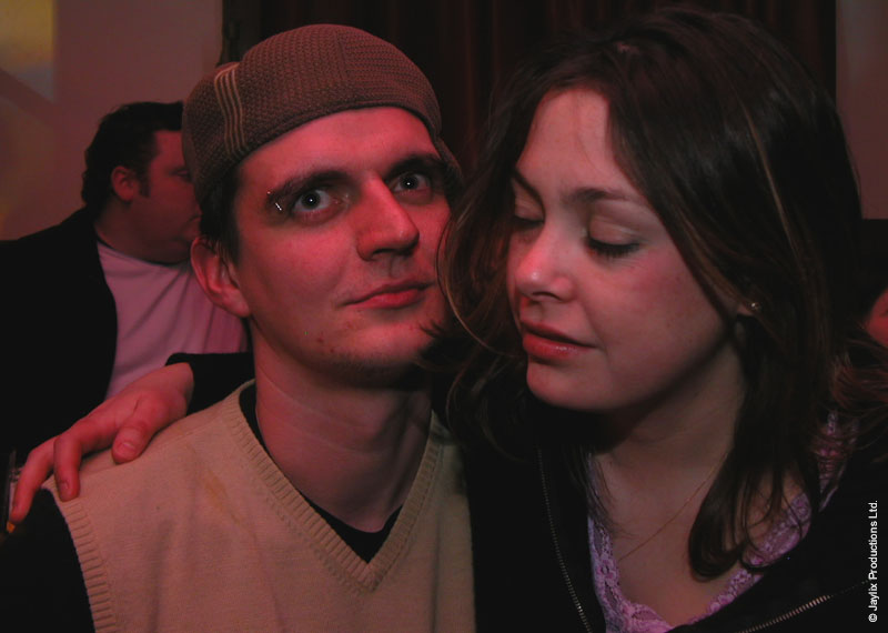 A man in a flat cap and a woman with her eyes closed lean together, with a man behind them