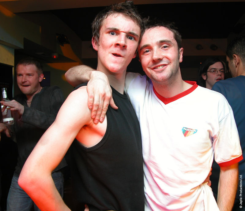 Two men pose with their arms around each other in a bar, one in a black vest and one in a white t-shirt