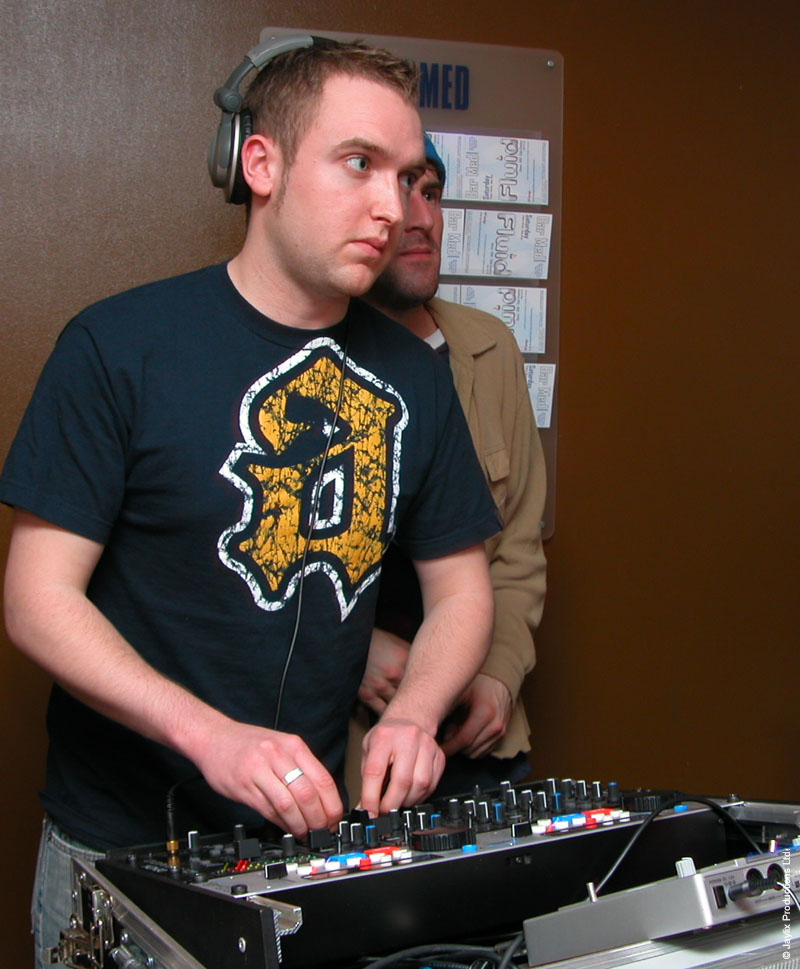 A DJ with headphones in a dark t-shirt mixes, with a person in a beige jacket behind and a Bar Med sign