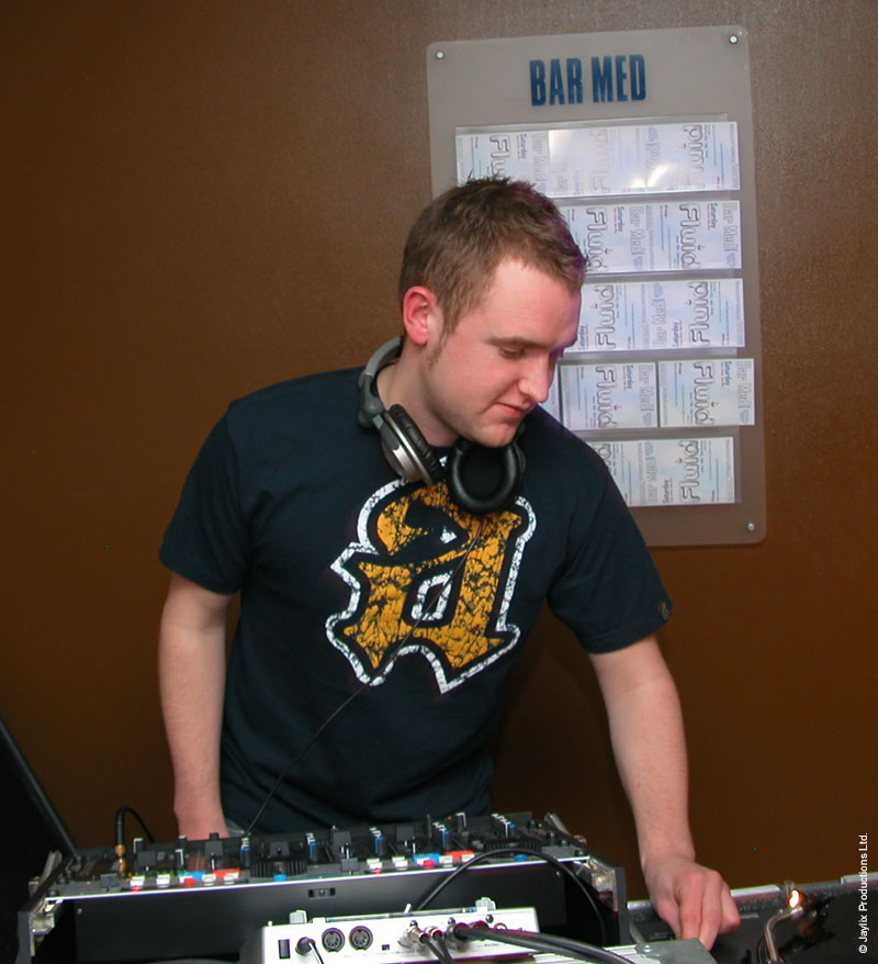 A DJ with headphones around his neck mixes at a desk in front of a Bar Med sign