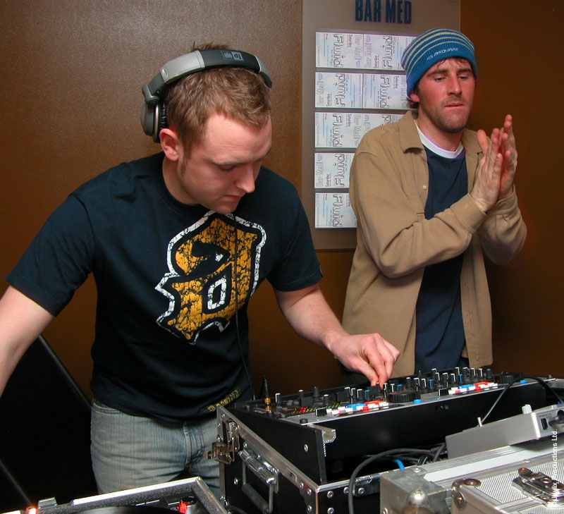 A DJ in headphones mixes while a person in a blue striped beanie claps beside him
