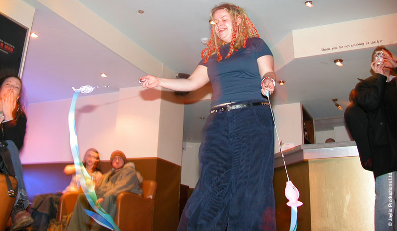 A person with curly hair dances in a bar holding two long ribbons