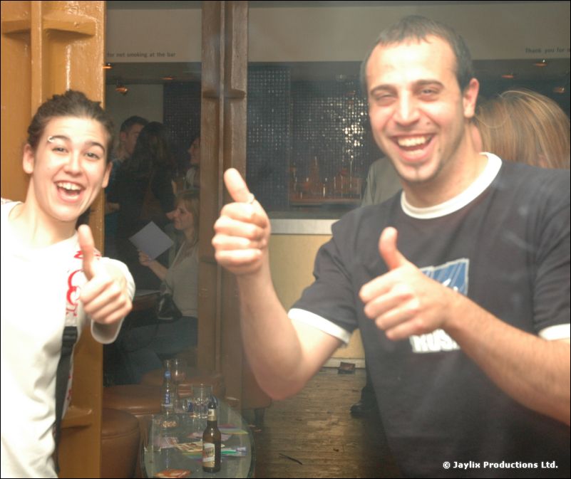 Two people laugh and give thumbs up in a bar, one in a black t-shirt and one in white