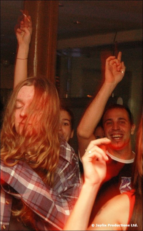 A person with long hair and a check shirt dances with eyes closed, a smiling man with raised arm behind