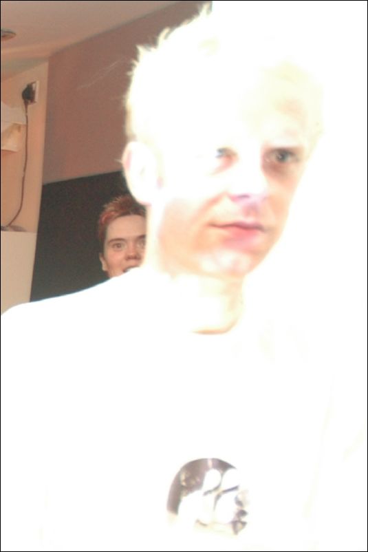 Overexposed image of a person with pale hair in a white top, another face behind
