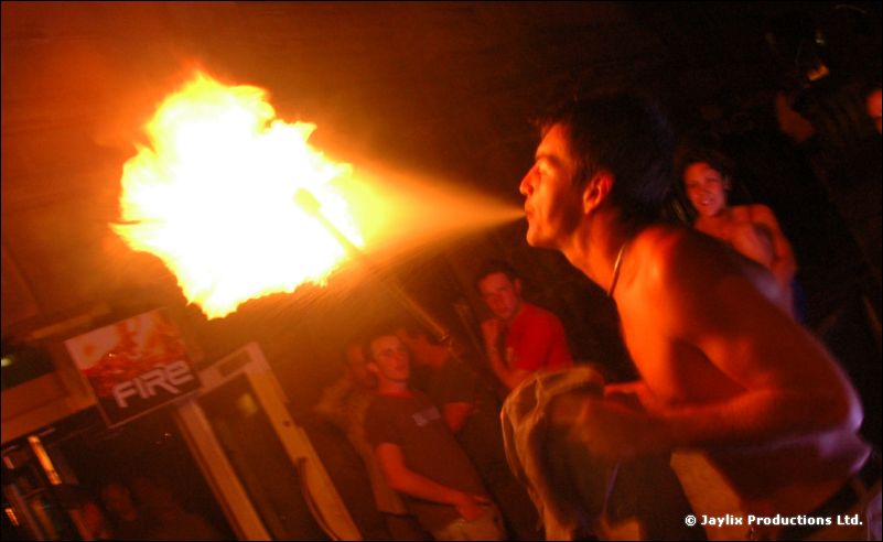 A shirtless man breathes a large ball of fire outdoors, with a FIRE sign behind