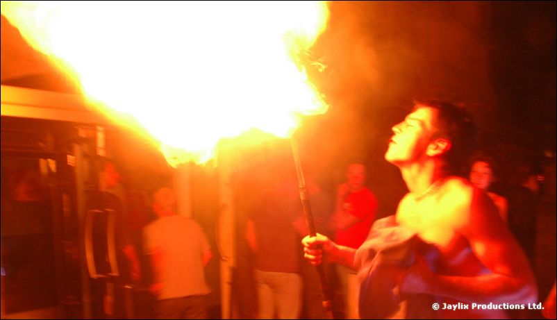 A shirtless man leans back holding a torch and blowing a large burst of fire