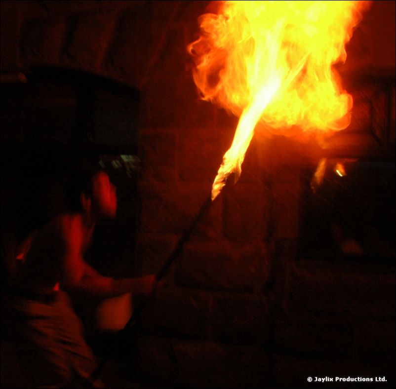 A large plume of fire from a torch beside a stone wall at night, a dark figure below