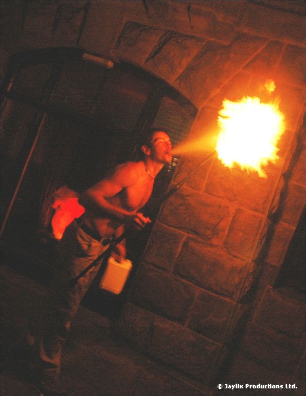 A shirtless man blows fire in a stone doorway, with a white container at his feet