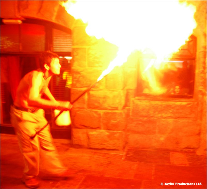 A person holding a torch breathes a large plume of fire beside a stone wall at night